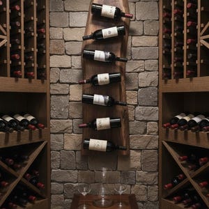 May include: A wine cellar with a rustic design. A wooden wine rack holds several bottles of red wine against a stone wall. Additional wine bottles are stored in wooden racks on either side. A small wooden table holds wine glasses and a decanter.