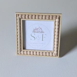 May include: A square light-colored wooden picture frame featuring a decorative beaded border. The center contains a white insert with a circular graphic of a house and the text S&F GENERATIONAL GOODS printed in a dark gray font.