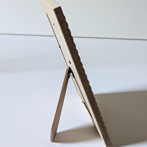 May include: A rectangular picture frame viewed from the side, featuring a light-colored wooden border with a textured, scalloped edge. The frame is supported by a dark brown wooden easel-style stand attached to the back panel.