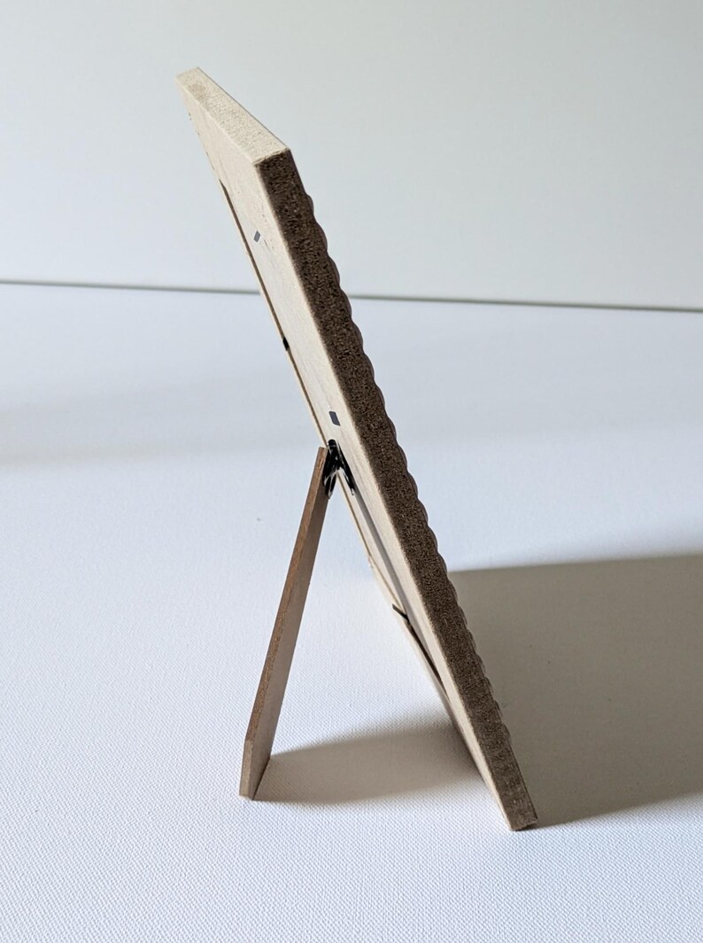 May include: A rectangular picture frame viewed from the side, featuring a light-colored wooden border with a textured, scalloped edge. The frame is supported by a dark brown wooden easel-style stand attached to the back panel.