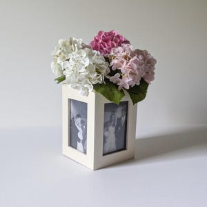 May include: A square white wooden photo frame vase featuring two visible vertical rectangular slots containing black and white photographs, topped with a cluster of artificial white, light pink, and dark pink hydrangea flowers with green leaves.