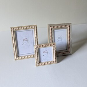 May include: Three picture frames with light wood finishes and beaded detailing along the inner edges. Each frame contains a white insert featuring the text S&F and a small house illustration. The set includes two rectangular frames and one square frame.