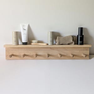 May include: A light-toned wooden wall shelf featuring seven rounded pegs along the bottom edge. The shelf holds two beige deodorant sticks, a white tube of toothpaste with a horse logo, a small wooden brush, a folded beige cloth, and a black bottle.