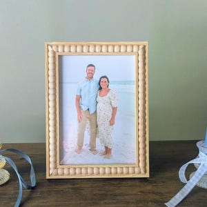 May include: A rectangular picture frame with a light-colored natural wood finish and a decorative beaded border. The frame holds a photograph of two people standing on a sandy beach, with the person on the left wearing a light blue shirt and tan pants.