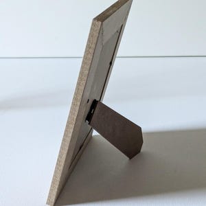 May include: A rectangular picture frame viewed from the side and back, featuring a light brown wood-grain finish. The frame includes a hinged support stand extending outward to allow the frame to stand upright on a flat surface.