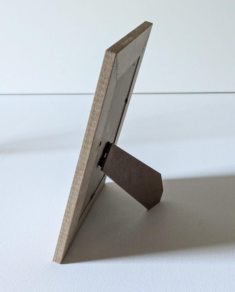 May include: A rectangular picture frame viewed from the side and back, featuring a light brown wood-grain finish. The frame includes a hinged support stand extending outward to allow the frame to stand upright on a flat surface.