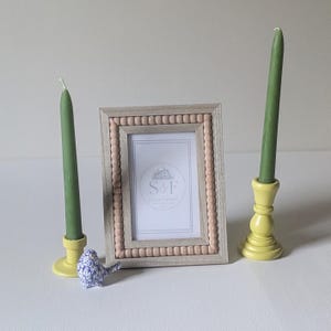 May include: A rectangular picture frame with a light wood finish and a border of small, round, light-colored wooden beads. Two tall, sage green taper candles stand in bright yellow candle holders. A small, blue and white speckled bird figurine.
