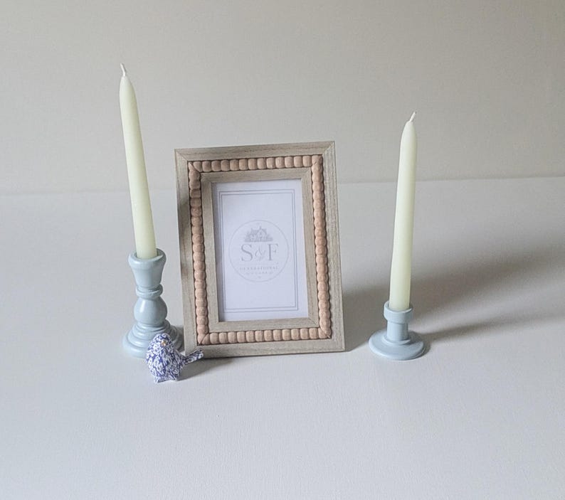 May include: A rectangular picture frame with a light wood finish and a beaded inner border, containing a white insert with the text S&F. Two light blue candle holders with white taper candles and a small blue and white patterned bird figurine.
