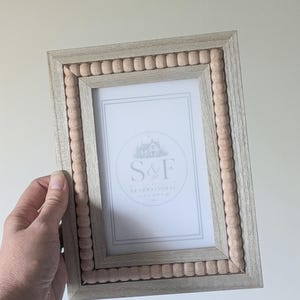 May include: A rectangular picture frame with a light gray wood grain finish and an inner border of small, round, light beige wooden beads. The center insert features a white background with the text S&F GENERATIONAL GOODS in a circular logo.