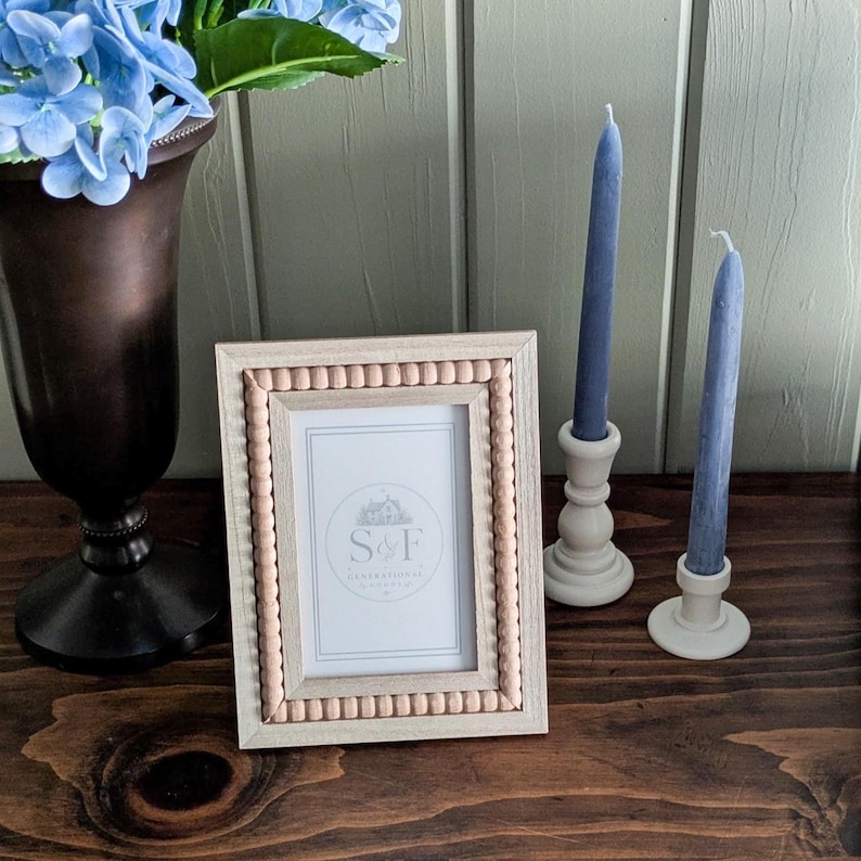 May include: A rectangular picture frame with a light wood finish and a border of small, round, light-colored wooden beads. The frame contains a white insert featuring the text S&F inside a circular logo with a house illustration.
