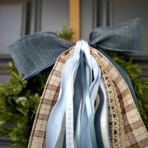 May include: A decorative bow featuring a wide blue denim-textured ribbon, layered with brown and white checkered fabric, light blue satin ribbons, and a strip of cream-colored lace trim. The ribbons are arranged in a cascading, multi-textured cluster.