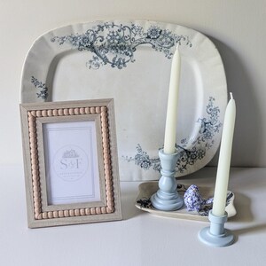 May include: A rectangular picture frame with a light wood finish and a beaded inner border, containing a white insert with the text S&F. Behind it sits a large oval white platter with blue floral patterns, two light blue candle holders, and a small blue bird figurine.