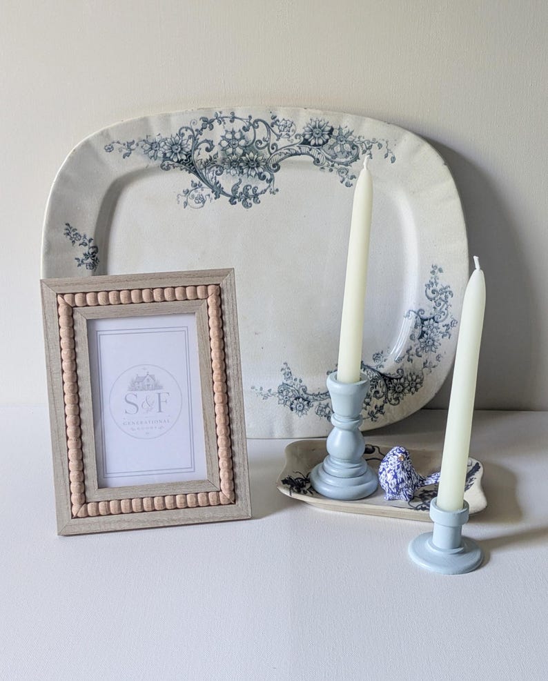 May include: A rectangular picture frame with a light wood finish and a beaded inner border, containing a white insert with the text S&F. Behind it sits a large oval white platter with blue floral patterns, two light blue candle holders, and a small blue bird figurine.