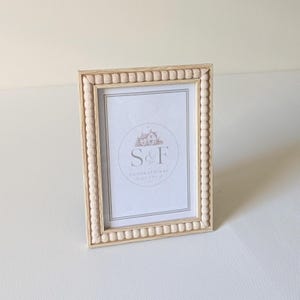 May include: A rectangular light wood picture frame featuring a decorative beaded border. The frame contains a white insert with a circular logo displaying a house illustration and the text S&F GENERATIONAL WORKS in gray lettering.