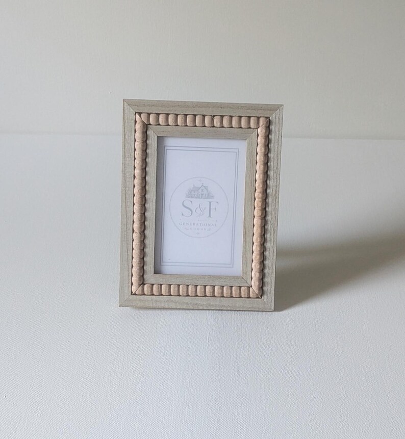 May include: A rectangular picture frame with a light wood finish and a border of small, round, light-colored wooden beads. The frame contains a white insert featuring a circular logo with the text S&F and a small house illustration.