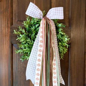 May include: A round green boxwood wreath adorned with a large bow made of light brown and white gingham ribbon. Several long, hanging ribbon streamers in various textures and patterns, including orange pumpkin prints, solid velvet, and gingham, drape down.