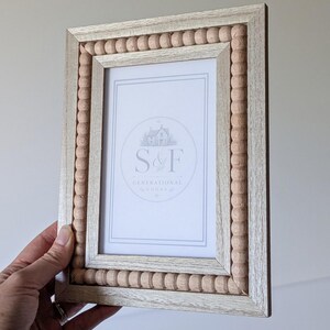 May include: A rectangular picture frame with a light wood finish and a border of small, natural-colored wooden beads. The center insert features a white background with the text S&F GENERATIONAL GOODS and a small house illustration.