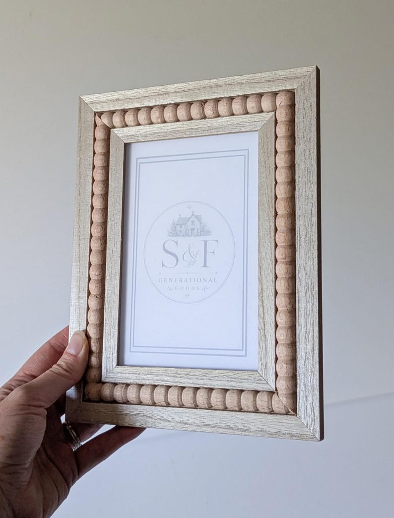 May include: A rectangular picture frame with a light wood finish and a border of small, natural-colored wooden beads. The center insert features a white background with the text S&F GENERATIONAL GOODS and a small house illustration.