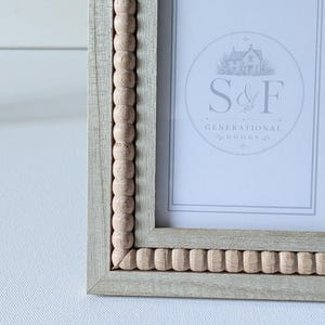 May include: A light gray wooden picture frame featuring a decorative inner border of small, natural-colored wooden beads. The frame contains a white insert with a circular logo displaying a house illustration and the text S&F GENERATIONAL GOODS.