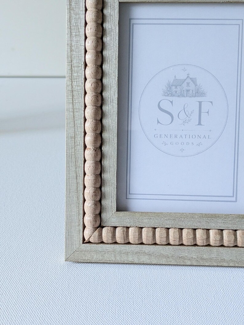 May include: A light gray wooden picture frame featuring a decorative inner border of small, natural-colored wooden beads. The frame contains a white insert with a circular logo displaying a house illustration and the text S&F GENERATIONAL GOODS.