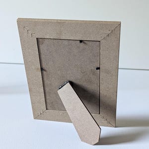 May include: A light brown rectangular picture frame made of composite wood material, viewed from the back. The frame features a hinged support stand and four corner joints decorated with etched arrow-like patterns.