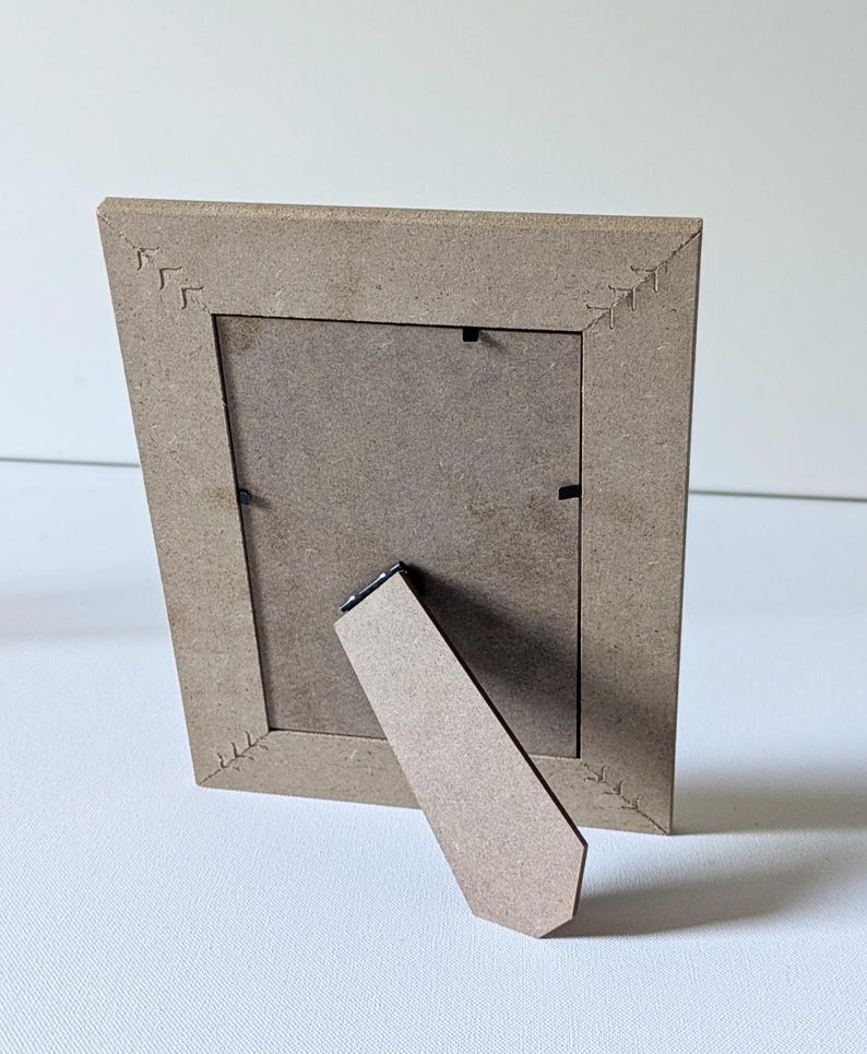May include: A light brown rectangular picture frame made of composite wood material, viewed from the back. The frame features a hinged support stand and four corner joints decorated with etched arrow-like patterns.