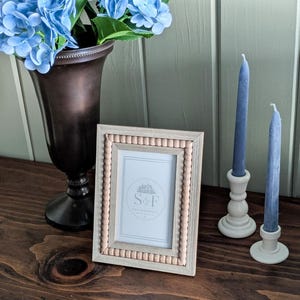 May include: A rectangular picture frame with a light wood finish and a beaded inner border. The frame contains a white insert with the text S&F. Beside it are two blue taper candles in cream-colored holders and a dark bronze vase with blue hydrangeas.