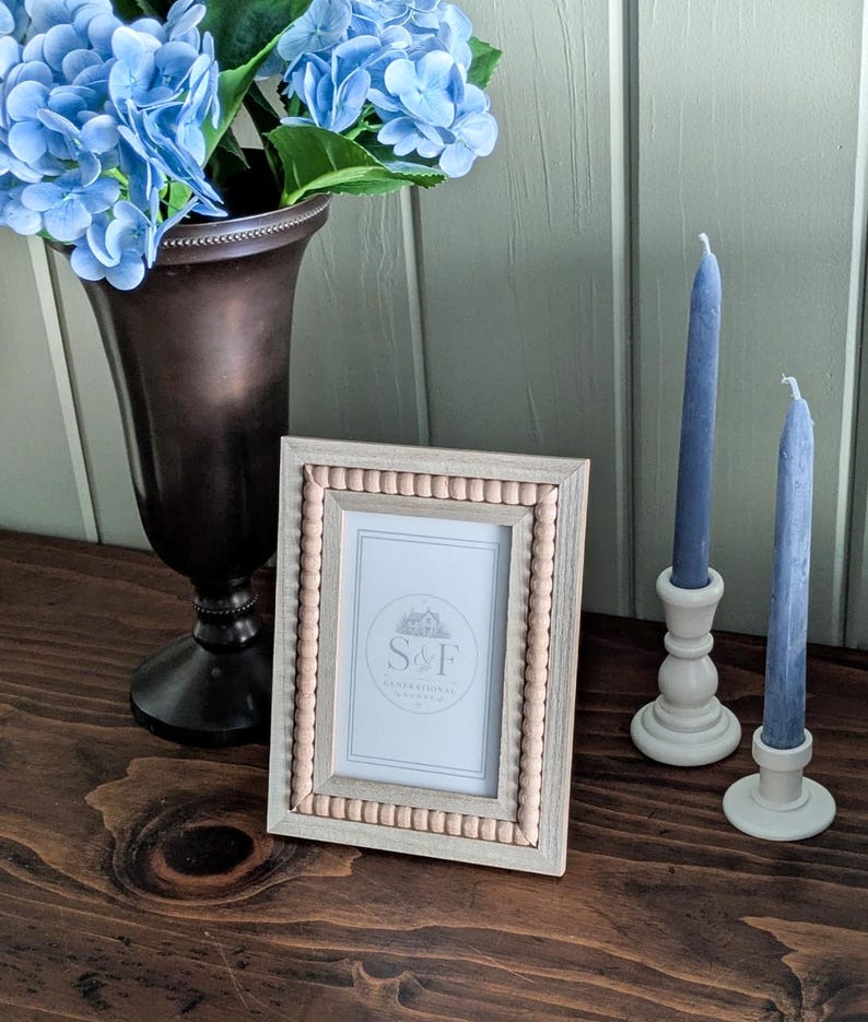 May include: A rectangular picture frame with a light wood finish and a beaded inner border. The frame contains a white insert with the text S&F. Beside it are two blue taper candles in cream-colored holders and a dark bronze vase with blue hydrangeas.