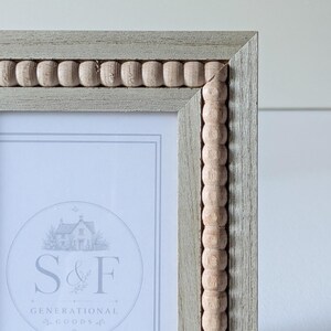 May include: A rectangular picture frame with a light gray wood grain finish and a decorative inner border of small, natural-colored wooden beads. The frame contains a white insert with the text S&F GENERATIONAL GOODS.