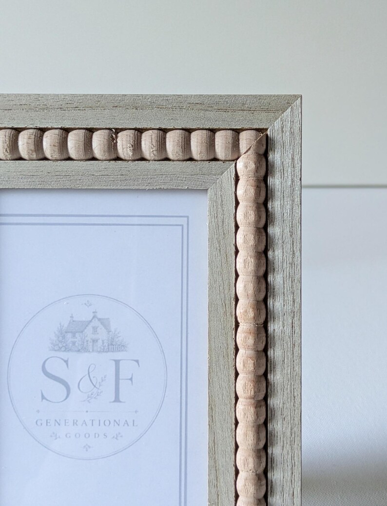 May include: A rectangular picture frame with a light gray wood grain finish and a decorative inner border of small, natural-colored wooden beads. The frame contains a white insert with the text S&F GENERATIONAL GOODS.