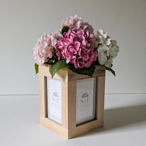 May include: A square light-colored wooden frame with four sides, each containing a white insert featuring a circular logo with the text S&F. A large bouquet of artificial hydrangea flowers in shades of light pink, dark pink, and white sits on top.