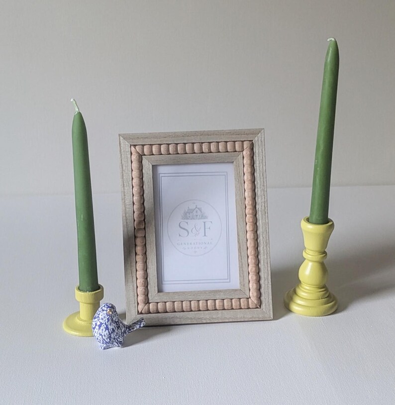 May include: A rectangular picture frame with a light wood finish and a beaded inner border, containing a white insert with the text S&F. Two tall green taper candles in yellow candle holders flank the frame, with a small blue bird figurine.