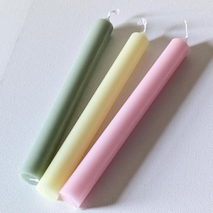 May include: Three smooth, cylindrical taper candles arranged diagonally. The candles are colored in muted sage green, pale yellow, and soft pastel pink. Each candle features a small, white cotton wick at the top end and a flat base at the bottom.