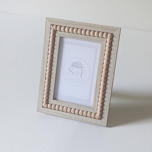 May include: A rectangular picture frame with a light gray wood grain finish and a border of small, round, light beige wooden beads. The frame contains a white insert featuring a circular logo with the text S&F GENERATIONAL.