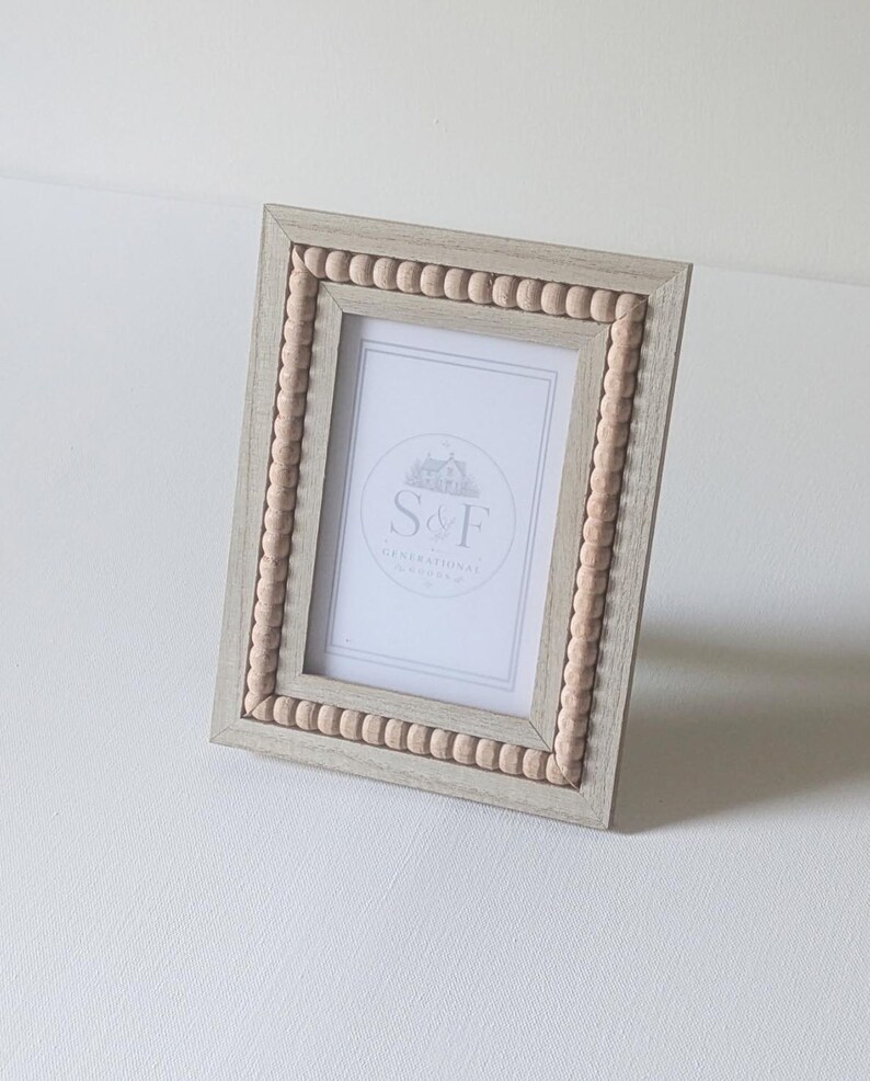 May include: A rectangular picture frame with a light gray wood grain finish and a border of small, round, light beige wooden beads. The frame contains a white insert featuring a circular logo with the text S&F GENERATIONAL.