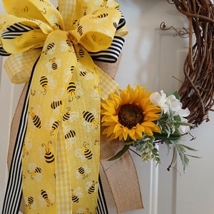 May include: A grapevine wreath adorned with a large yellow bow featuring a bee pattern, black and white stripes, and gingham. A sunflower and white flowers with greenery add a natural touch to this decorative wreath.