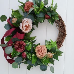 May include: A burgundy and blush floral wreath with greenery and a burgundy ribbon. The wreath is made of grapevine and is decorated with artificial flowers and leaves.