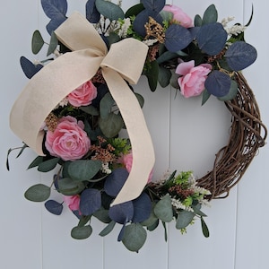May include: A decorative wreath featuring pink roses, dark blue eucalyptus leaves, and a beige ribbon bow. The wreath is made of woven brown twigs and is suitable for indoor or outdoor use.