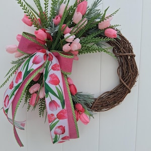 May include: A spring wreath featuring pink tulips and greenery arranged on a natural grapevine base. A pink ribbon with a tulip pattern and green polka dot trim adds a decorative touch to the floral arrangement.