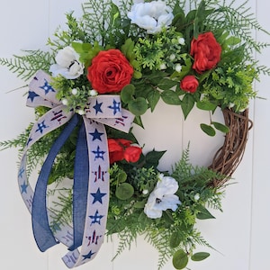 May include: A decorative wreath featuring red roses, white flowers, and green foliage arranged on a brown grapevine base. A blue and tan ribbon with star accents adds a patriotic touch. The wreath is hanging on a white door.