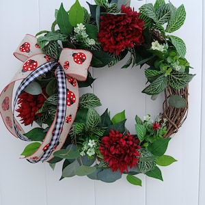 May include: A red and green wreath with a black and white ladybug ribbon. The wreath is made of twigs and has red flowers and green leaves.