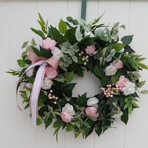 May include: A circular wreath with a mix of greenery, pink and white flowers, and small white berries. A pink ribbon and white ribbon hang from the top. The wreath is hanging on a white door.