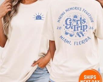 May include: A cream-coloured t-shirt featuring a small blue sun graphic on the front left chest. The back displays a large blue circular design with the text "Girls Trip," "Making Memories Together," "Miami, Florida," "2026," palm trees, sunglasses, and flip-flops.