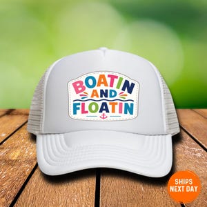 May include: A white trucker hat featuring a central patch with the colorful text "BOATIN AND FLOATIN" in a mix of pink, orange, green, and blue letters, accented by small decorative wave lines and a small pink anchor icon centered at the bottom.