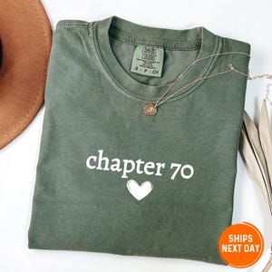 May include: An olive green crewneck t-shirt featuring the white text "chapter 70" above a white heart outline. The shirt has a Comfort Colors brand label at the inner neckline and is displayed with a delicate gold-toned necklace with a small pendant.