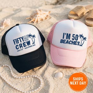 May include: Two trucker hats with white fronts and mesh backs rest on sand. One is black with "FIFTYAF CREW" and palm tree graphics. The other is pink with "I'M 50 BEACHES" and palm tree graphics. Both hats have text and palm tree graphics in navy blue.