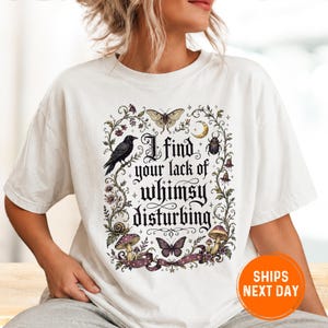 May include: A white short-sleeve t-shirt featuring a central graphic with the text I find your lack of whimsy disturbing. The design includes a black raven, a moth, a beetle, a snail, mushrooms, and a crescent moon, all framed by floral vines.