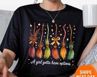 May include: Black t-shirt featuring a colorful illustration of six witch brooms with various designs and bows. The text "A girl gotta have options" is printed below the brooms. The shirt is a casual wear item.
