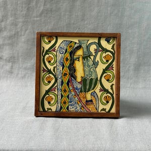 May include: Square ceramic tile art in a wooden frame. The tile features a profile of a person with a blue and yellow head covering, holding a blue and green jug. The background is yellow with green and orange floral designs.
