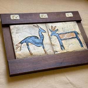 May include: A rectangular dark brown wooden frame holding two square ceramic tiles. Each tile features a dark blue stylised animal figure on a textured beige background. The top wooden rail includes three small, square, light-coloured ceramic inlays.